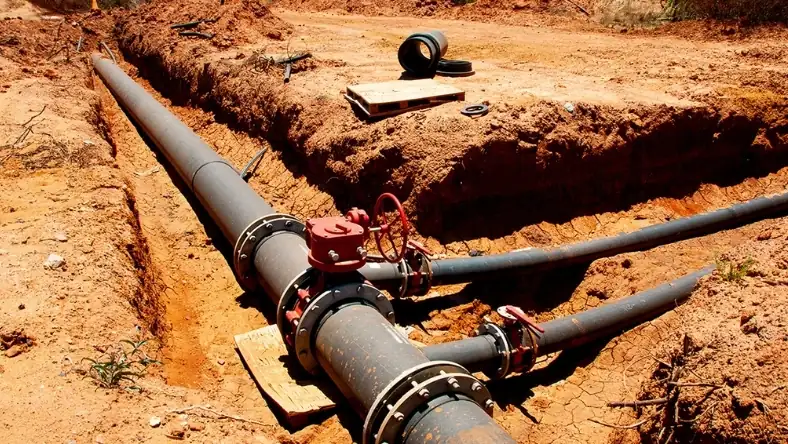 Steel pipeline fittings and red valve wheels visible in an open dirt trench during installation work