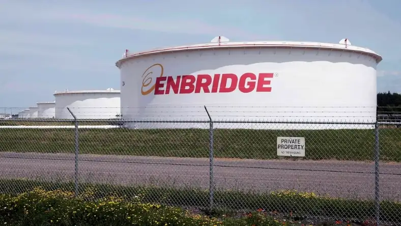 White oil storage tanks branded Enbridge behind a chain link fence with wildflowers in the foreground