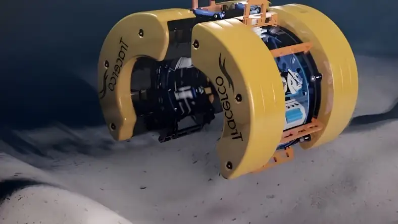 Yellow subsea robotic clamp device with orange fittings resting above a sandy rippled ocean floor bed